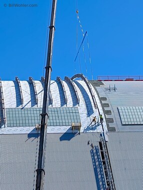 Packets of roof panels are hoisted in place and secured by climbers until they can be installed